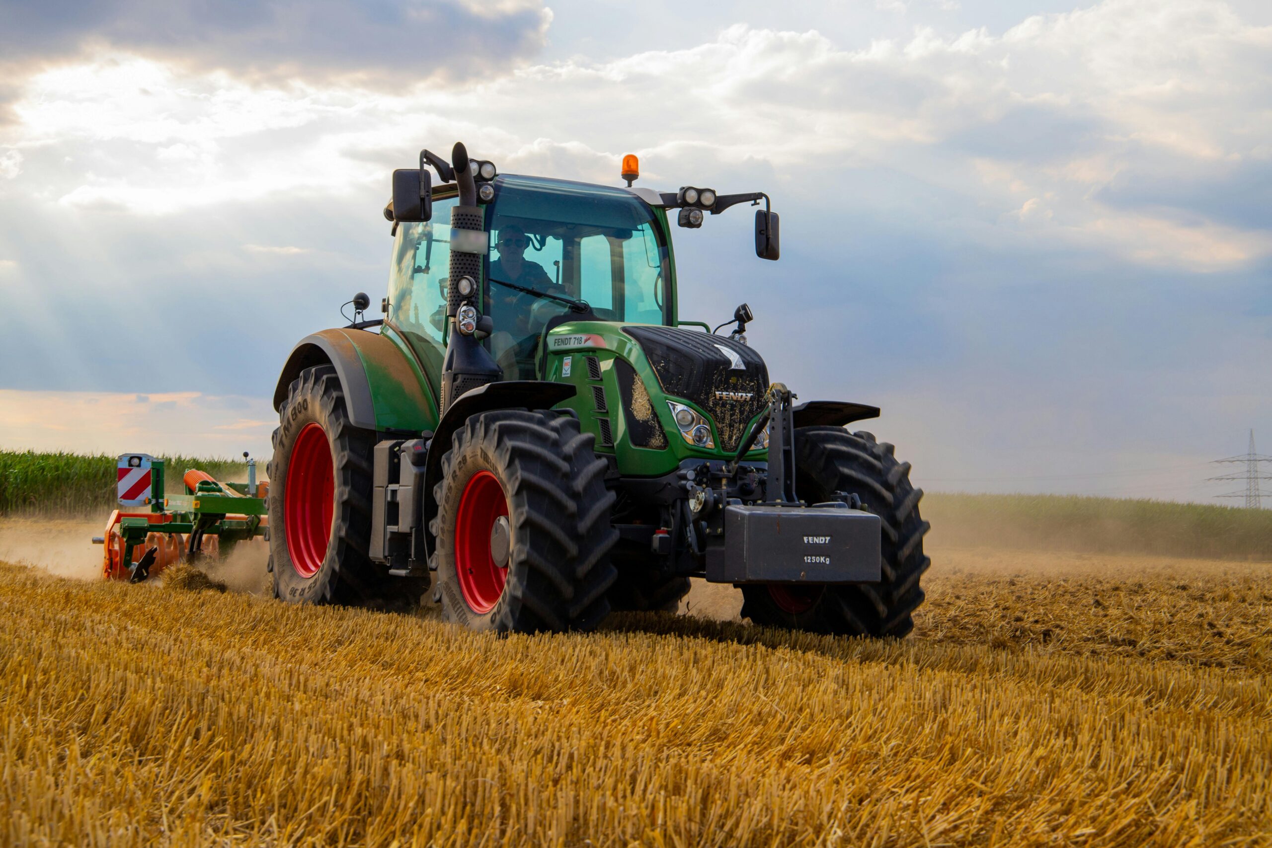 A powerful green tractor plowing a dusty wheat field under a cloudy summer sky in rural Germany.