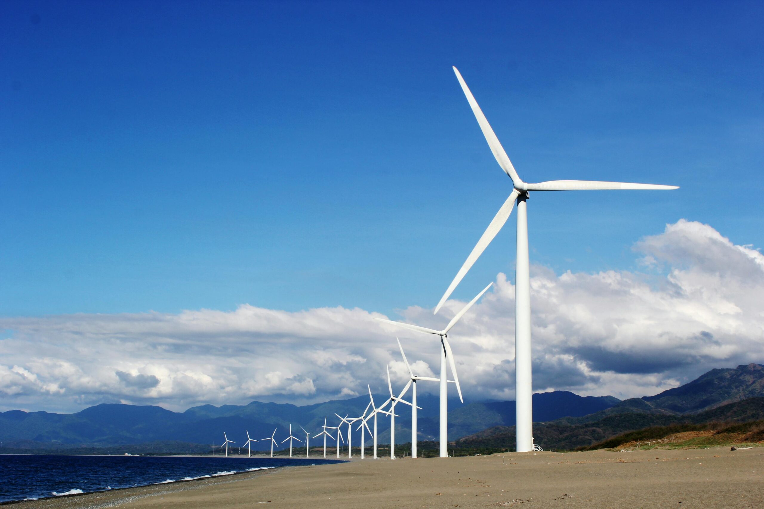 A row of wind turbines on a sandy coastline under a clear blue sky, promoting renewable energy.